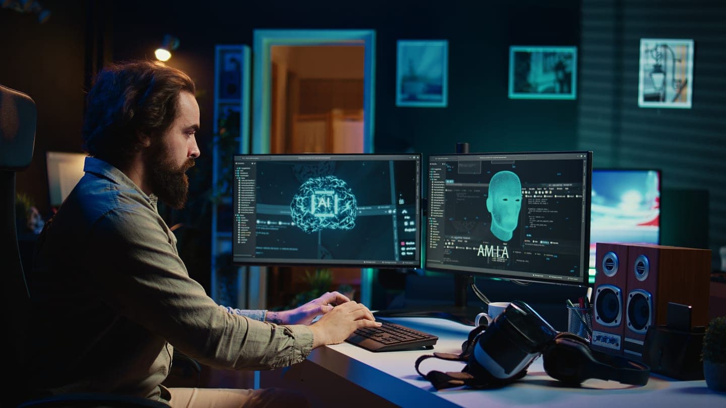 A bearded developer in a dark room types at two monitors showing a glowing cyan AI brain diagram and a digital face beside panels of code, with a VR headset on the desk.