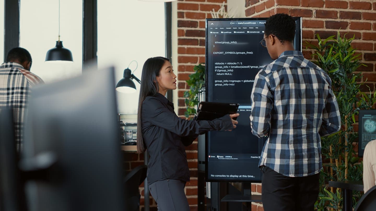 Two software engineers in a brick-walled office reviewing source code on a tall wall-mounted screen, one holding a tablet, while a colleague works at a desk