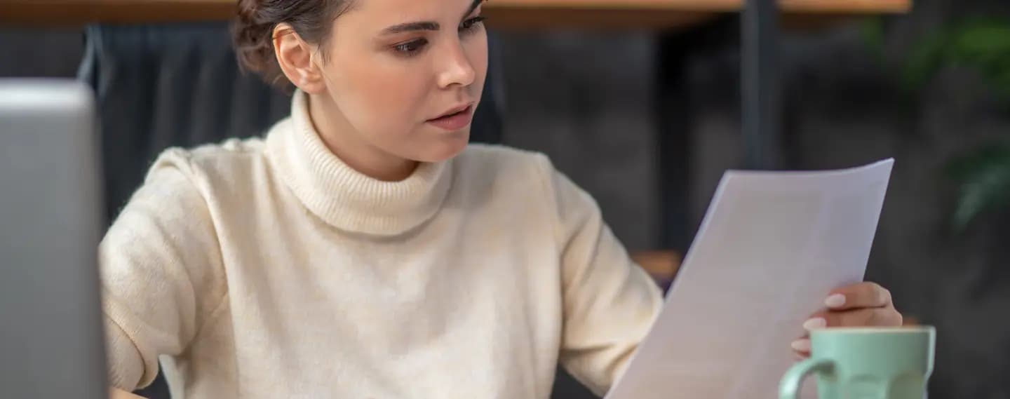 A woman in a cream turtleneck sits at a desk, reading a printed document held up in one hand with a concentrated, questioning expression, a laptop edge and a pale green mug beside her.