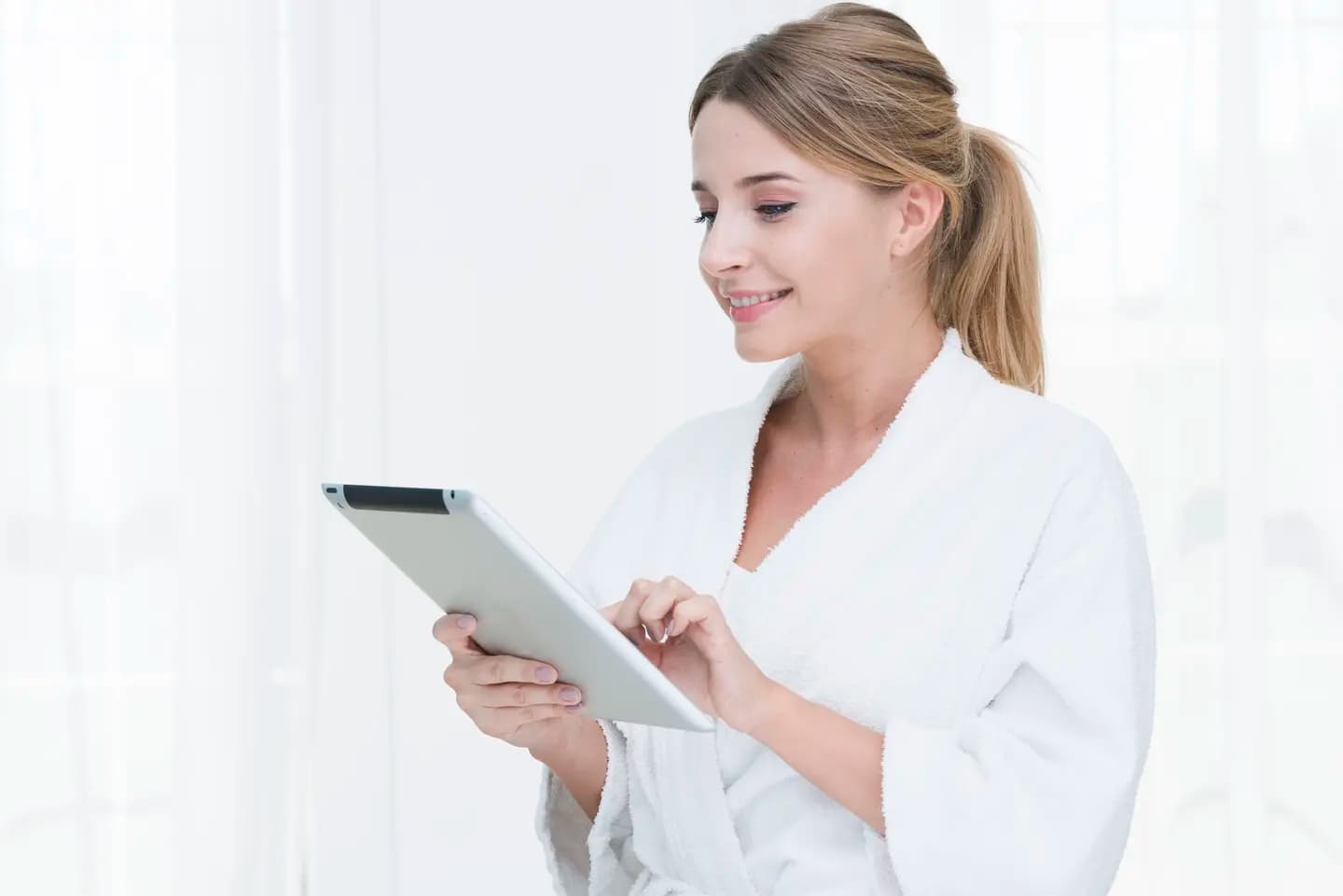 A woman in a white spa robe smiles at a tablet she holds in both hands, her ponytail over one shoulder against a bright window.