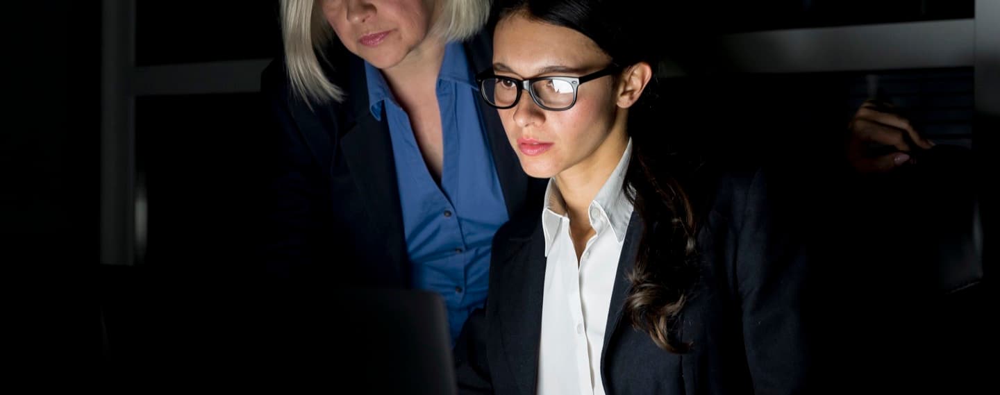 Two colleagues studying a laptop in a darkened office, their faces lit only by the screen.