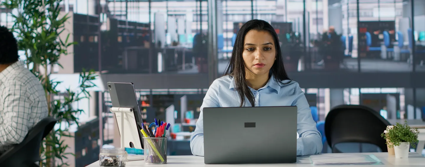A woman in a pale blue shirt works on a laptop at a desk in an open-plan office with glass partitions behind her.