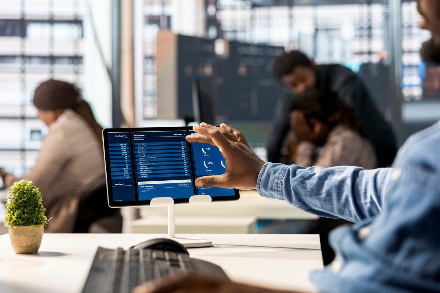 An analyst in a denim shirt taps a tablet showing a blue billing dashboard on a desk stand while colleagues work out of focus behind