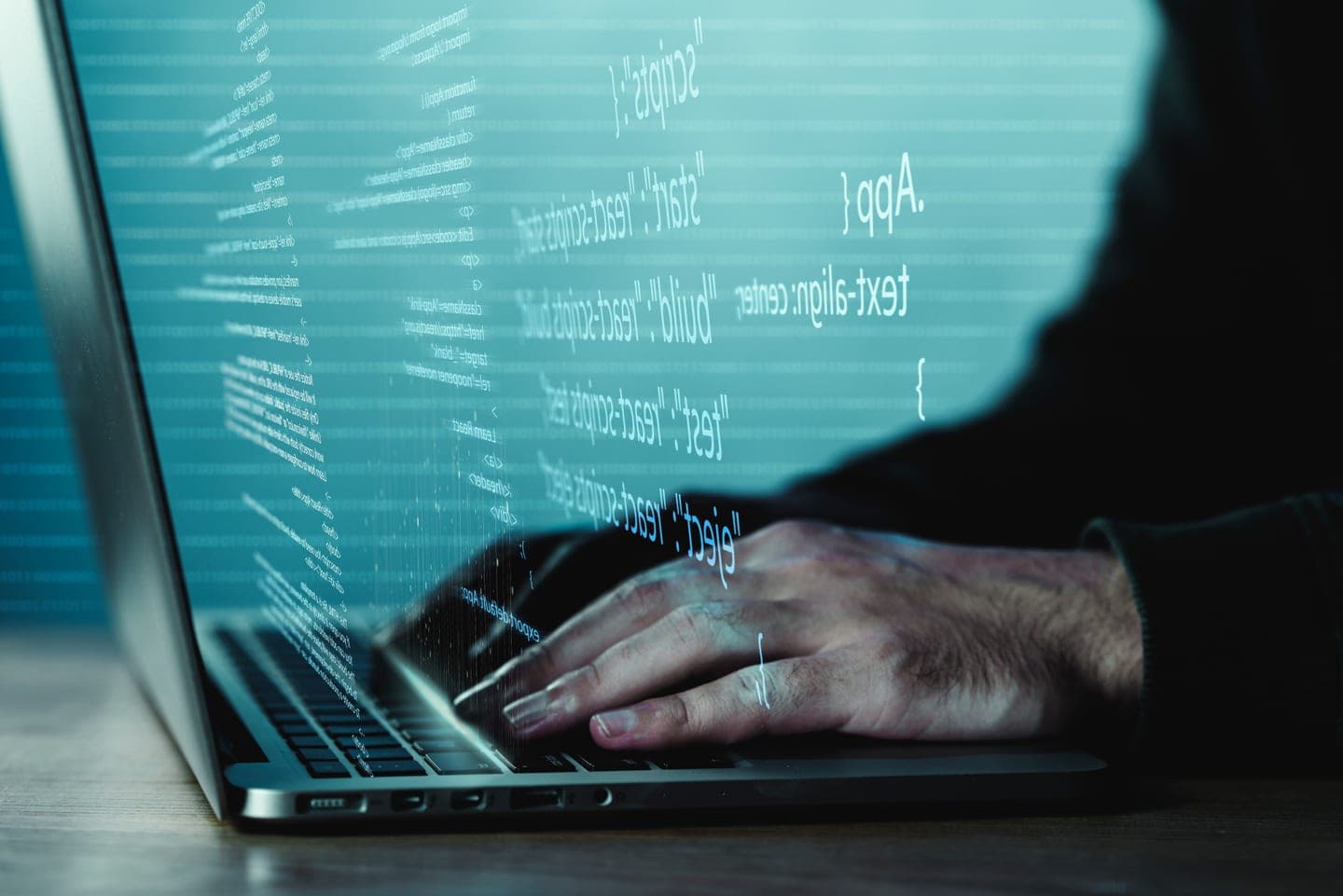 Hands typing on a laptop in a dark room with lines of code projected in glowing cyan across the screen and the coder's silhouette