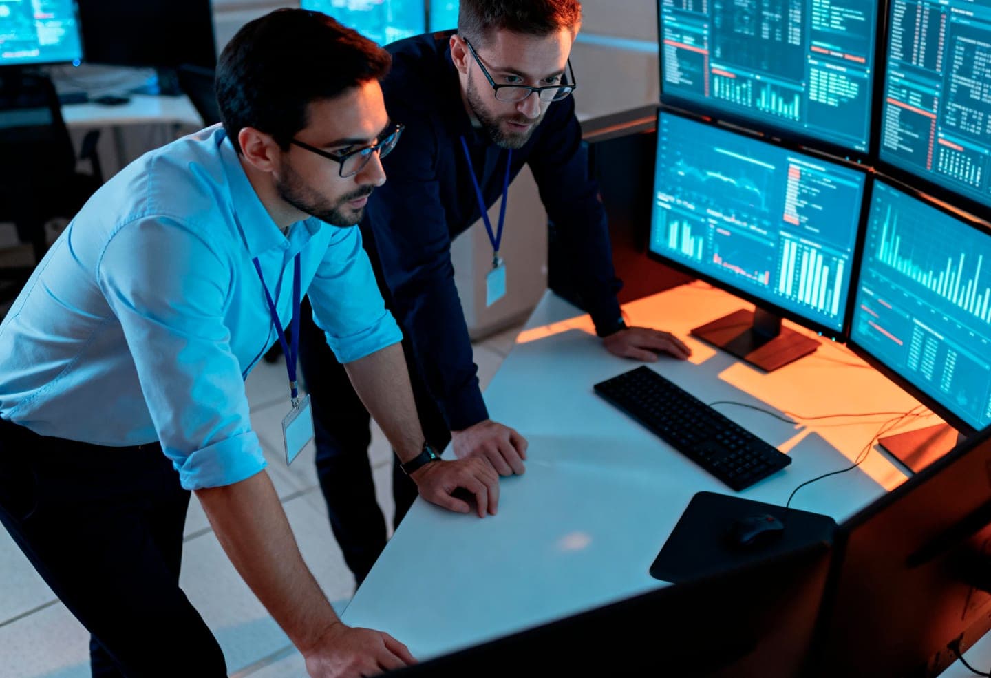Two engineers with lanyards lean over a desk in a dim control room, studying a wall of monitors filled with live charts and data panels in blue light
