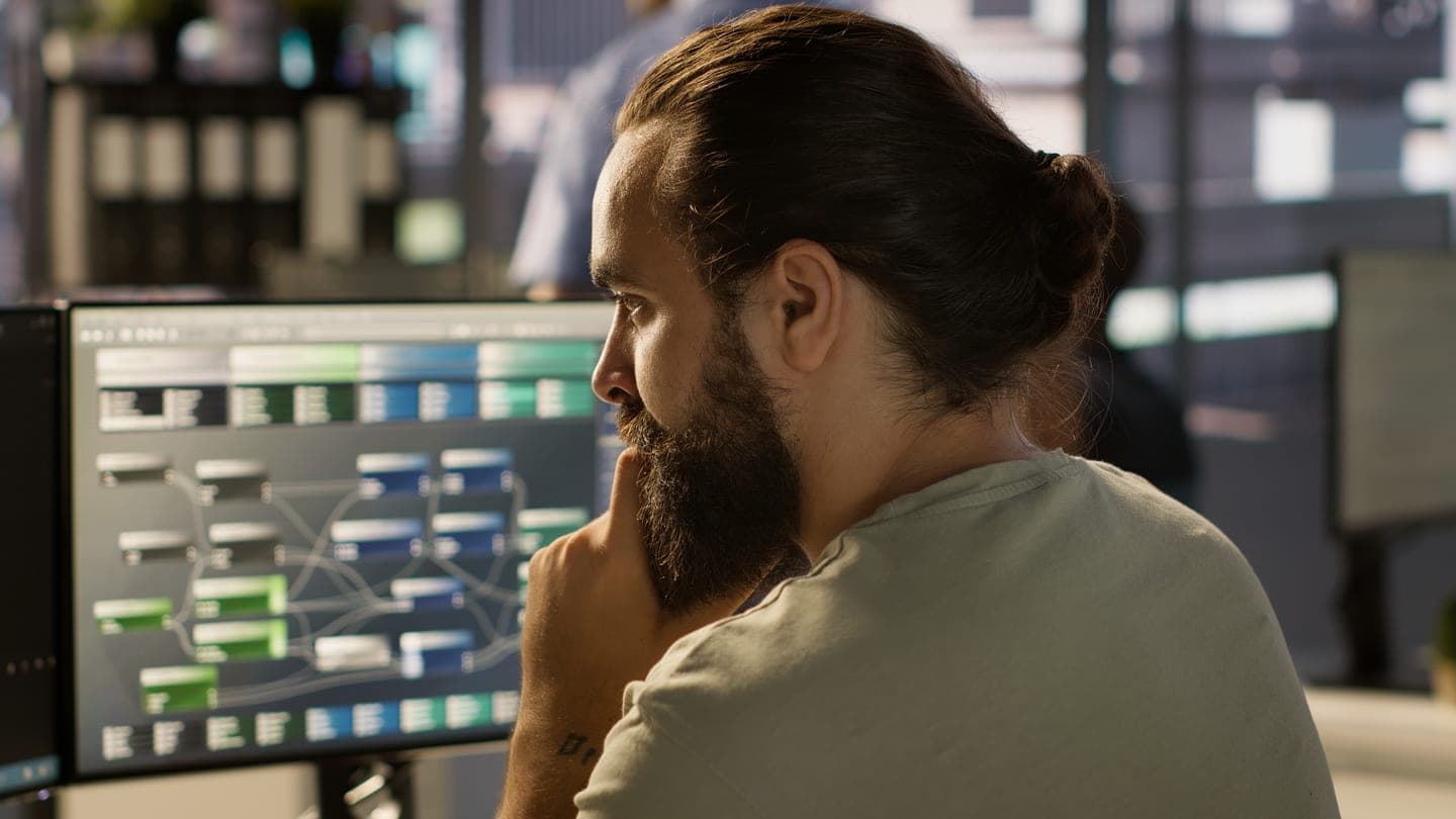 A bearded engineer with his hair tied back studies a monitor showing a node diagram of blue and green data pipeline blocks in a dim office