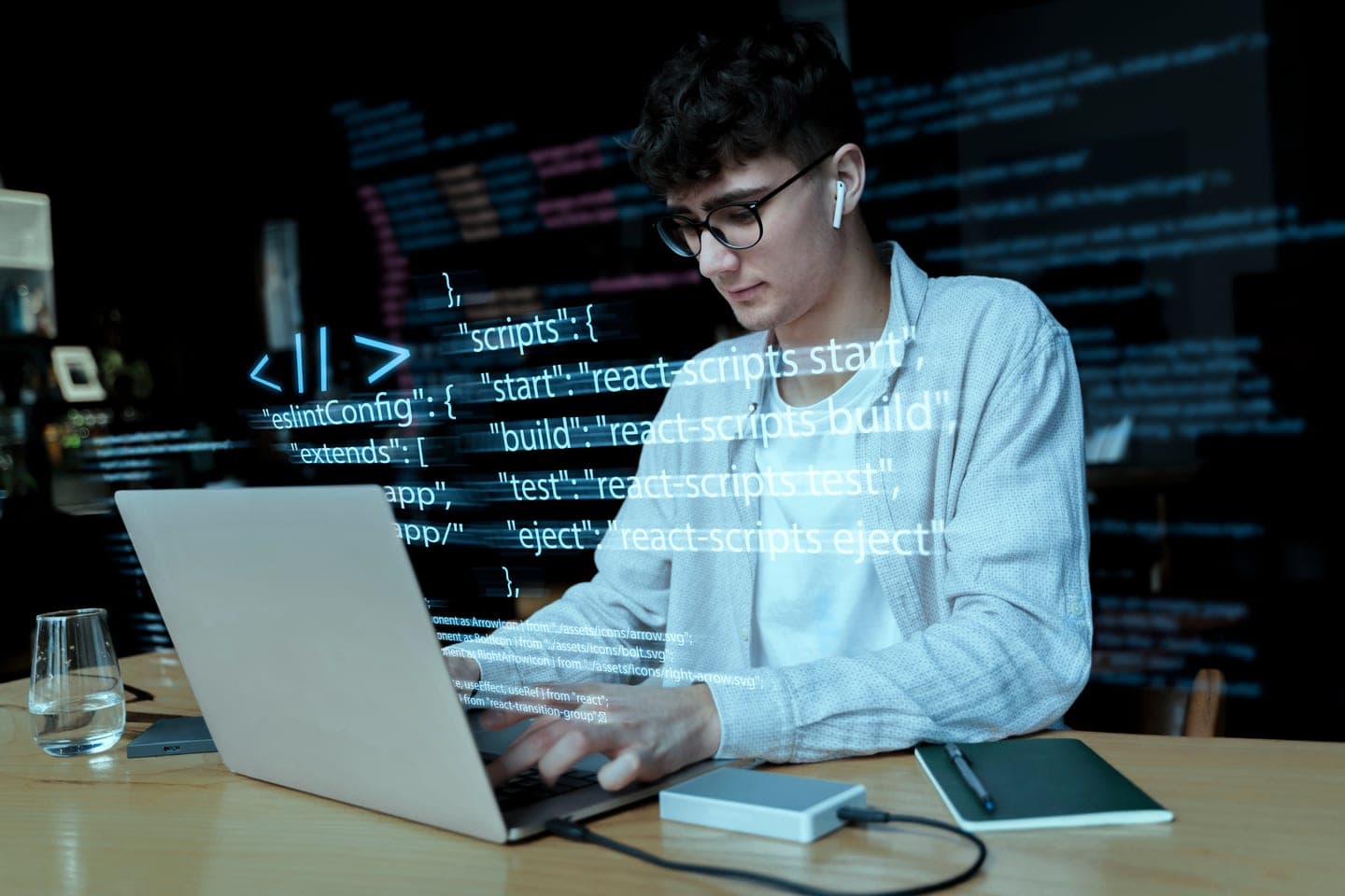 Developer in glasses typing on a laptop in a dark room while glowing lines of JSON configuration code float in the air in front of him