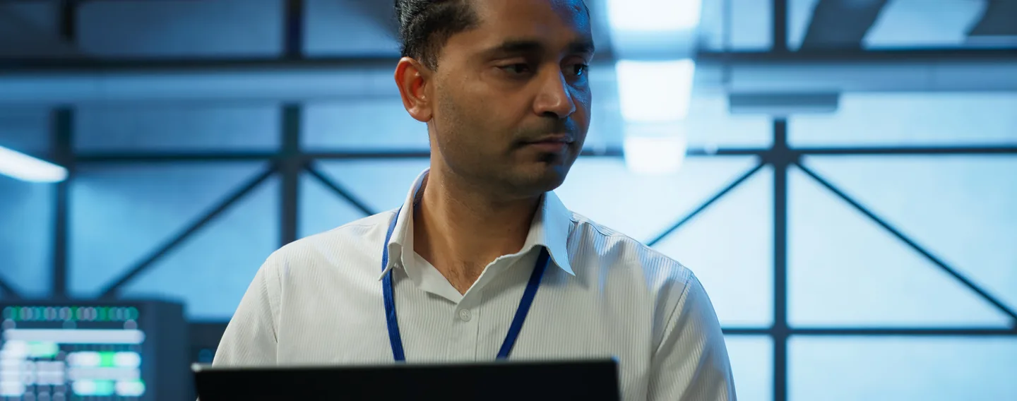 A man wearing a lanyard looks down at a laptop in a data centre, with server racks and steel roof framing behind him.