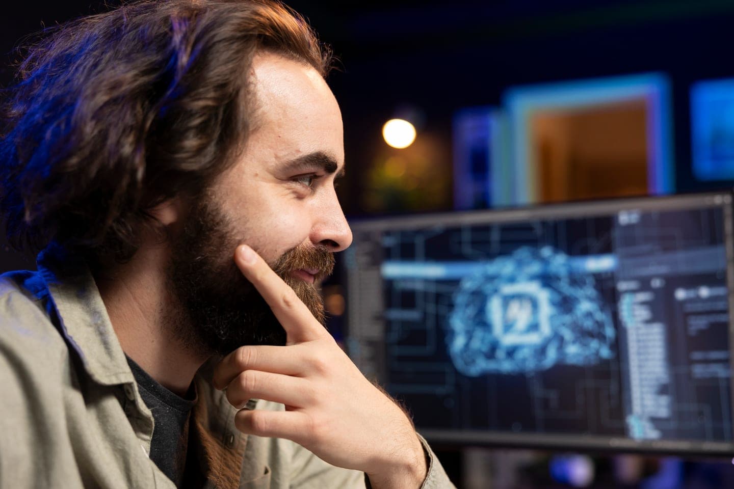 A bearded engineer with long hair rests his chin on his hand and smiles at a monitor in a dim room, where a glowing cyan neural network shaped like a brain sits among panels of code.