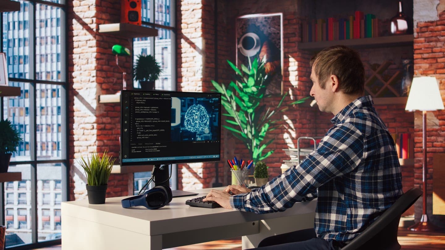 A developer in a plaid shirt types at a desk in a brick-walled home office, his monitor showing terminal code beside a glowing blue brain visualisation
