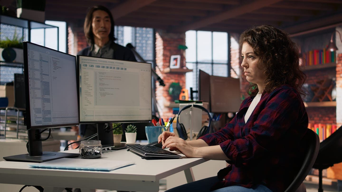 Developer in a checked shirt reviewing code on two monitors while a colleague stands behind her in a brick-walled office