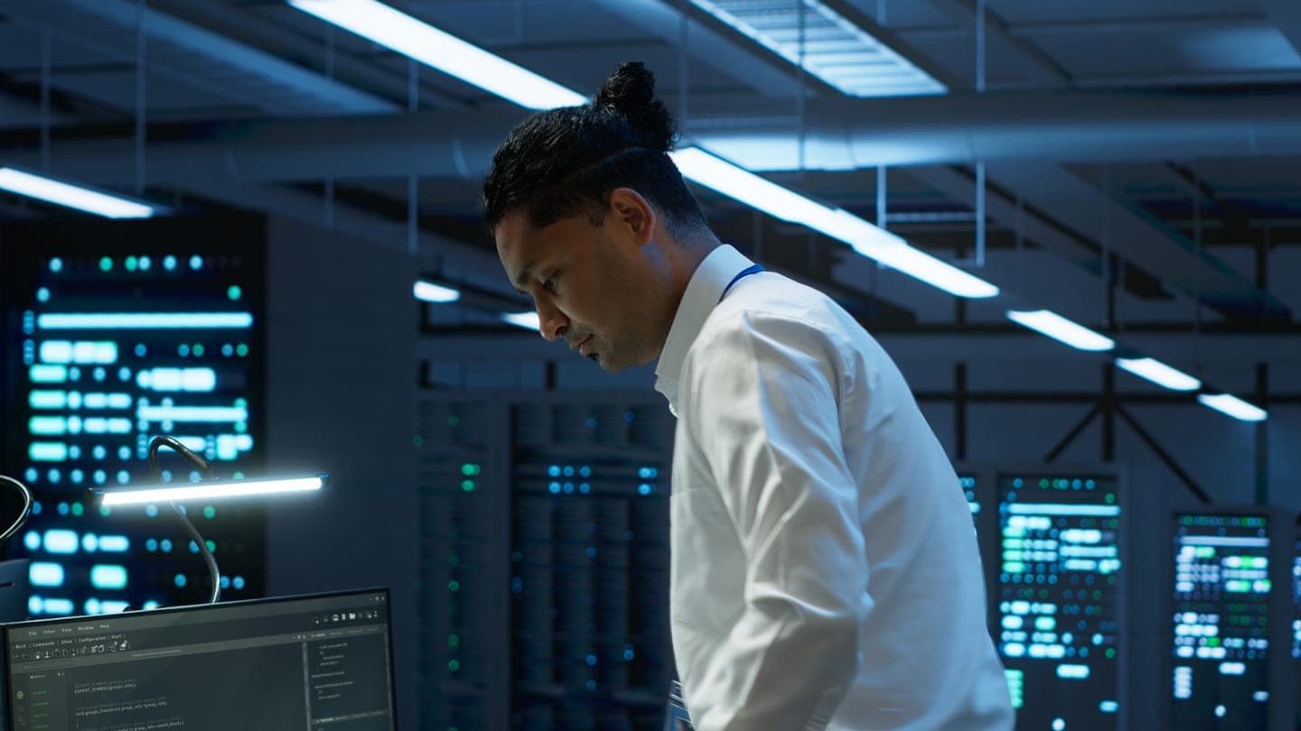 An engineer in a white shirt looks down at a terminal in a dim data centre, with rows of server racks and glowing green and blue status screens behind him.