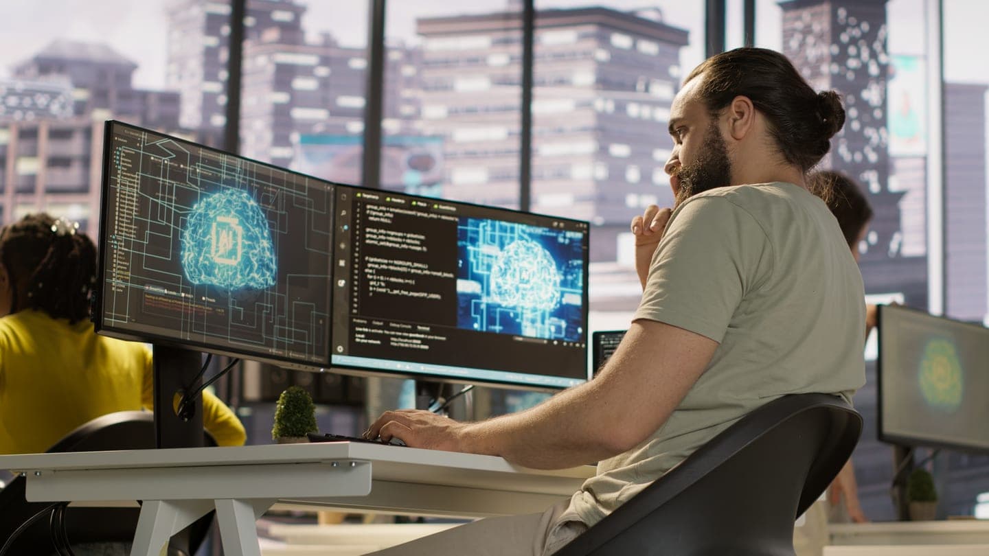 Engineer at a desk reviewing code beside two monitors that show a glowing blue AI chip brain inside a circuit board pattern