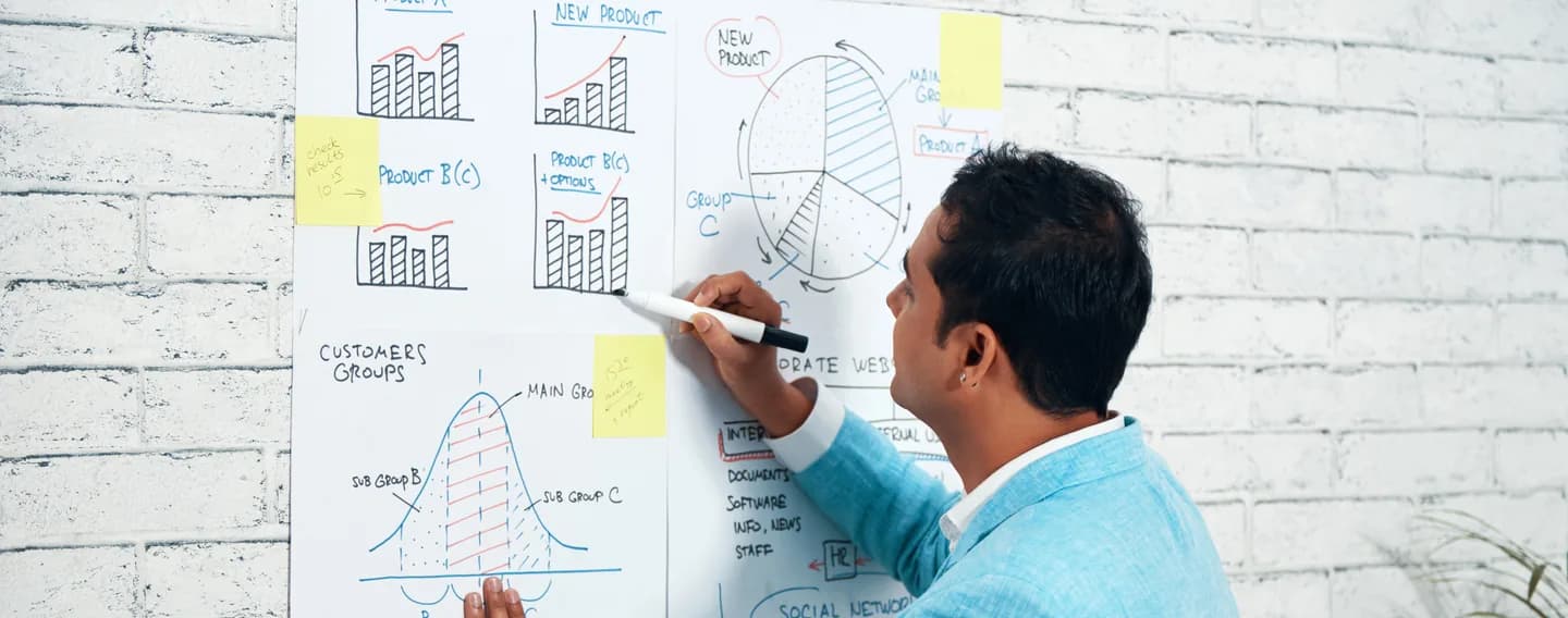 A man in a light blue jacket marks up hand-drawn charts and diagrams on paper sheets pinned to a white brick wall.