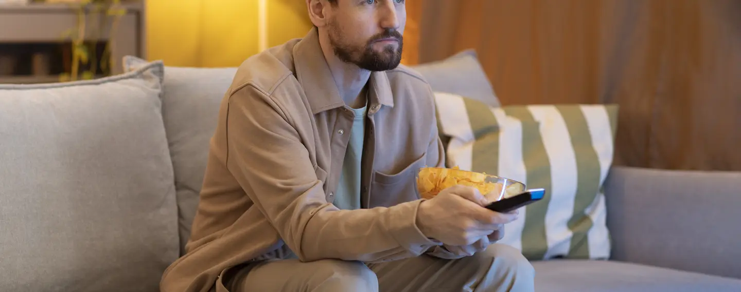 A bearded man in a tan jacket sits on a grey sofa in a warm-lit living room, holding a bowl of crisps in one hand and a TV remote control in the other.