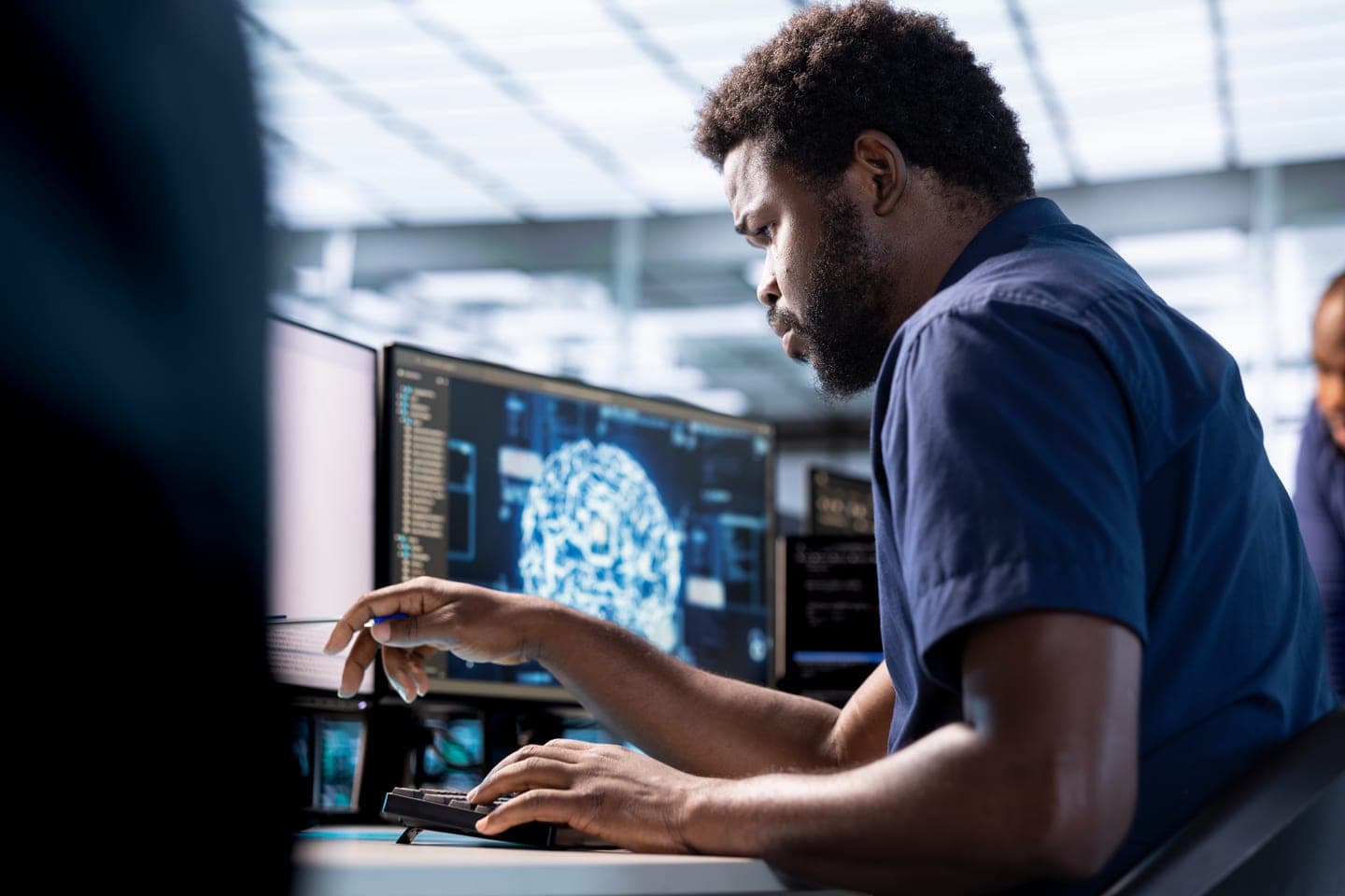 An engineer in a navy shirt points at a monitor showing a glowing blue brain graphic beside lines of code at a workstation in a bright server room