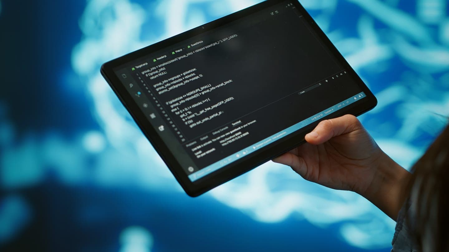 A hand holds a tablet showing lines of code in a dark editor, in front of a glowing cyan neural network pattern on a deep blue background