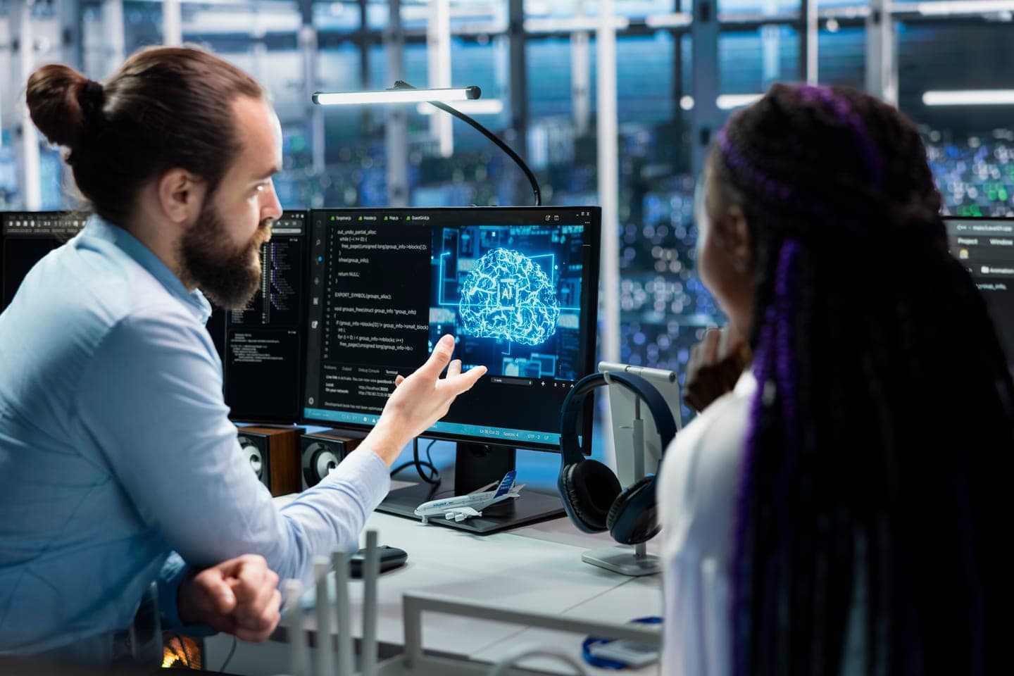 Two engineers in a data centre office discuss code and a glowing neural network model on a monitor, with server racks behind the glass