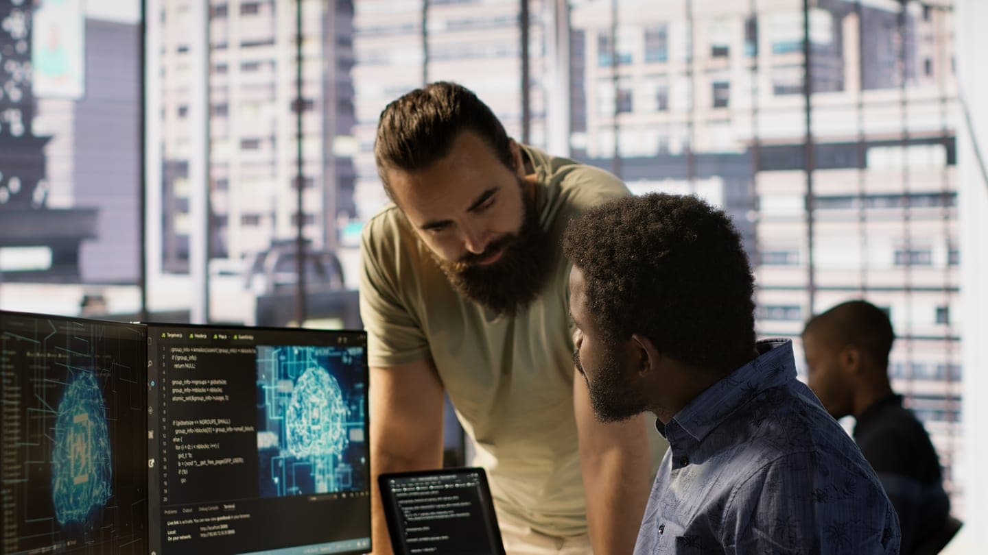 Manager leaning over a programmer at two monitors showing glowing blue AI neural-network models and code