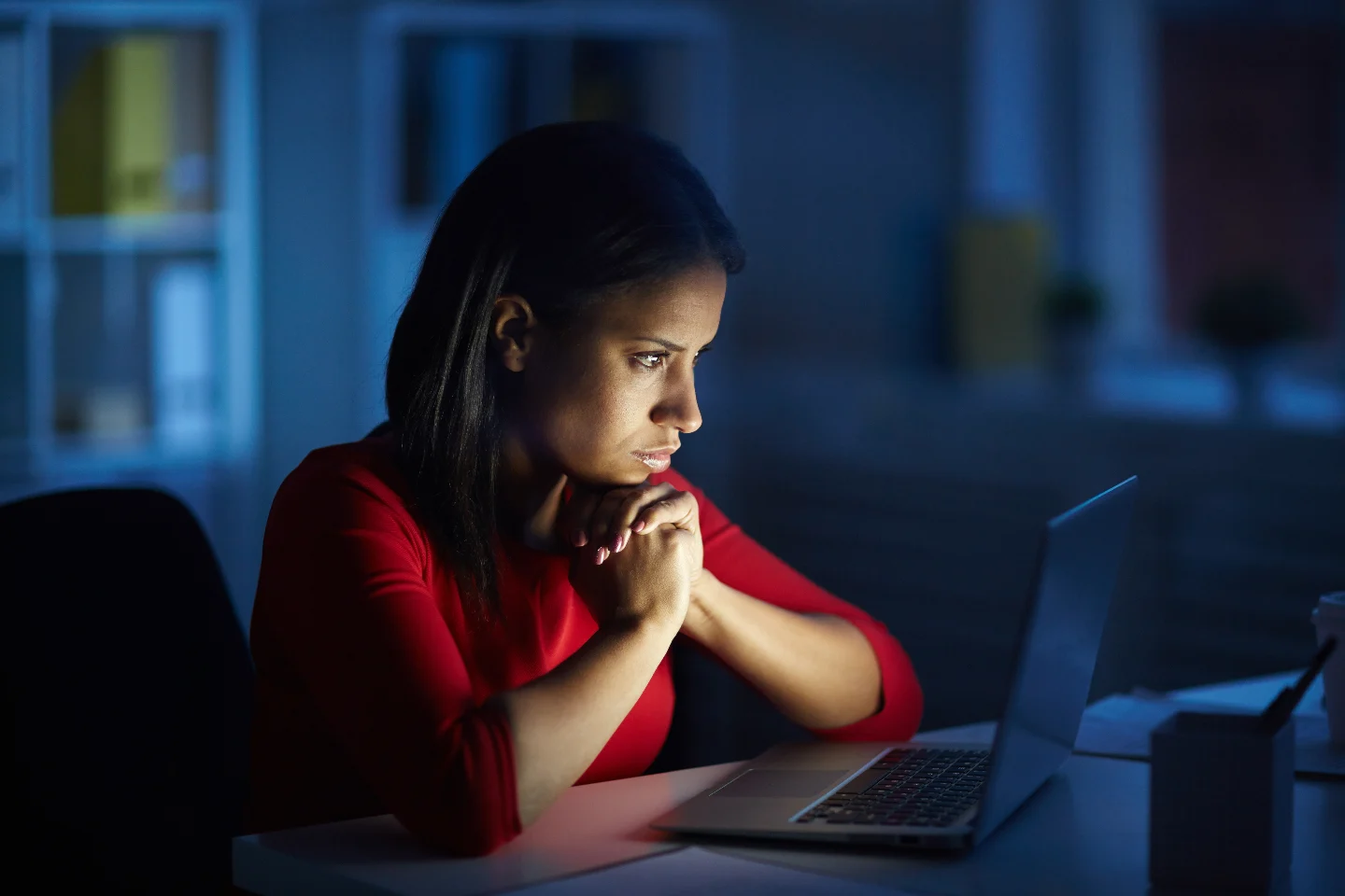 An engineer in a white shirt sits in a dimly lit server room, hand to chin, watching monitors that show a glowing blue neural network brain and lines of system code.