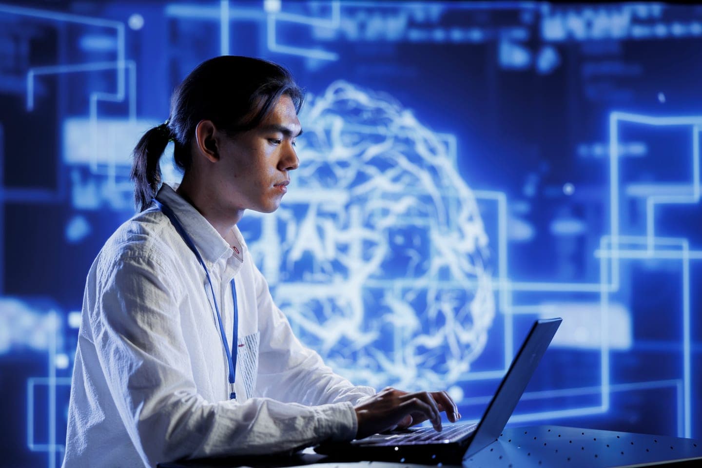 A young engineer in a white shirt types on a laptop in front of a large glowing blue display of a neural network brain and circuit lines in a dark data centre.