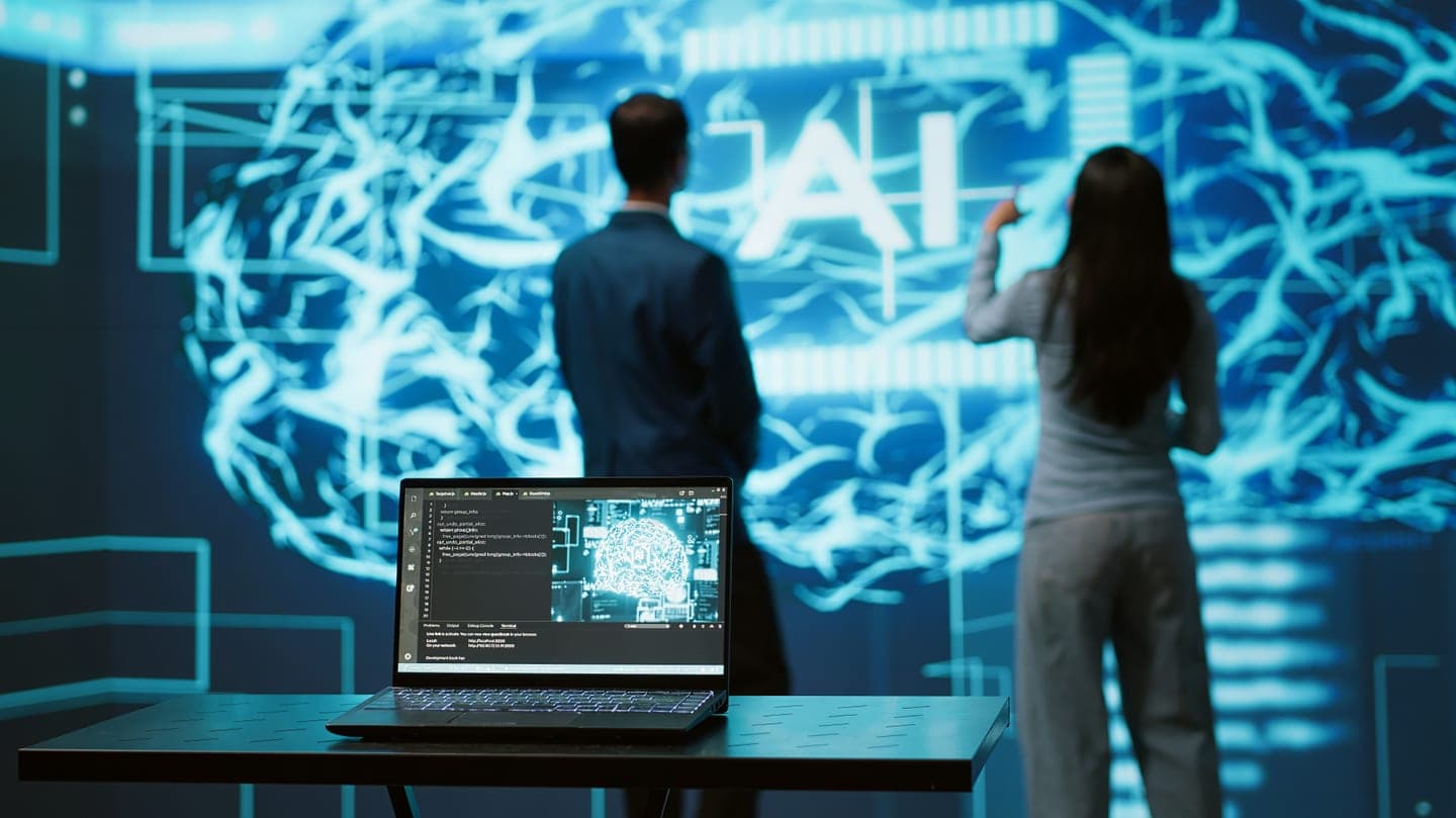 Two engineers stand before a wall-sized glowing cyan neural network display marked AI, with a laptop showing model code on a desk in front