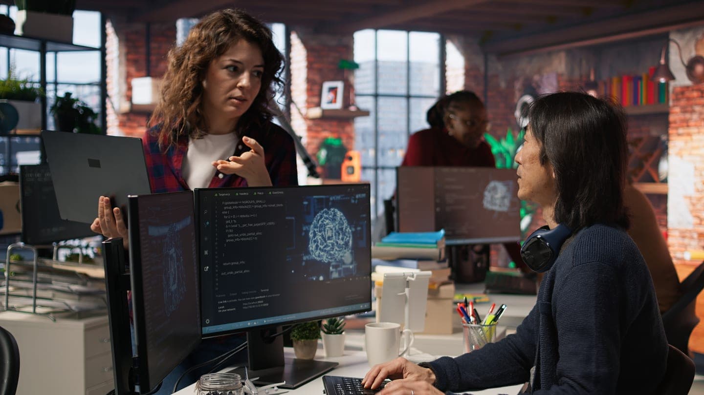 Woman holding a laptop asks a question of a developer whose monitors show code and a glowing AI brain graphic in a brick-walled office