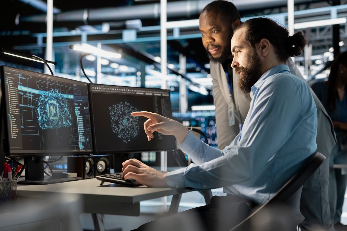 A bearded engineer in a light blue shirt points at a monitor showing a glowing blue neural network graphic while a colleague leans over his shoulder to review it, in a dimly lit server hall.