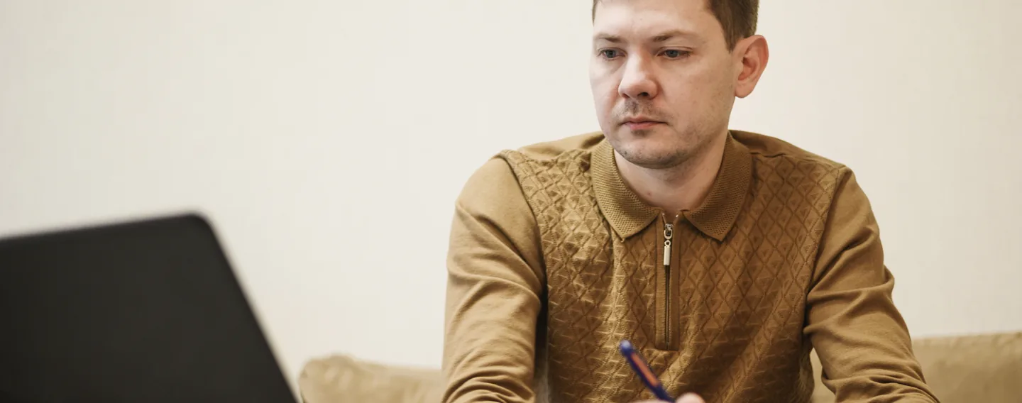 A man in a brown knitted shirt sits on a sofa holding a pen and looking down at his notes, with a laptop in the foreground.