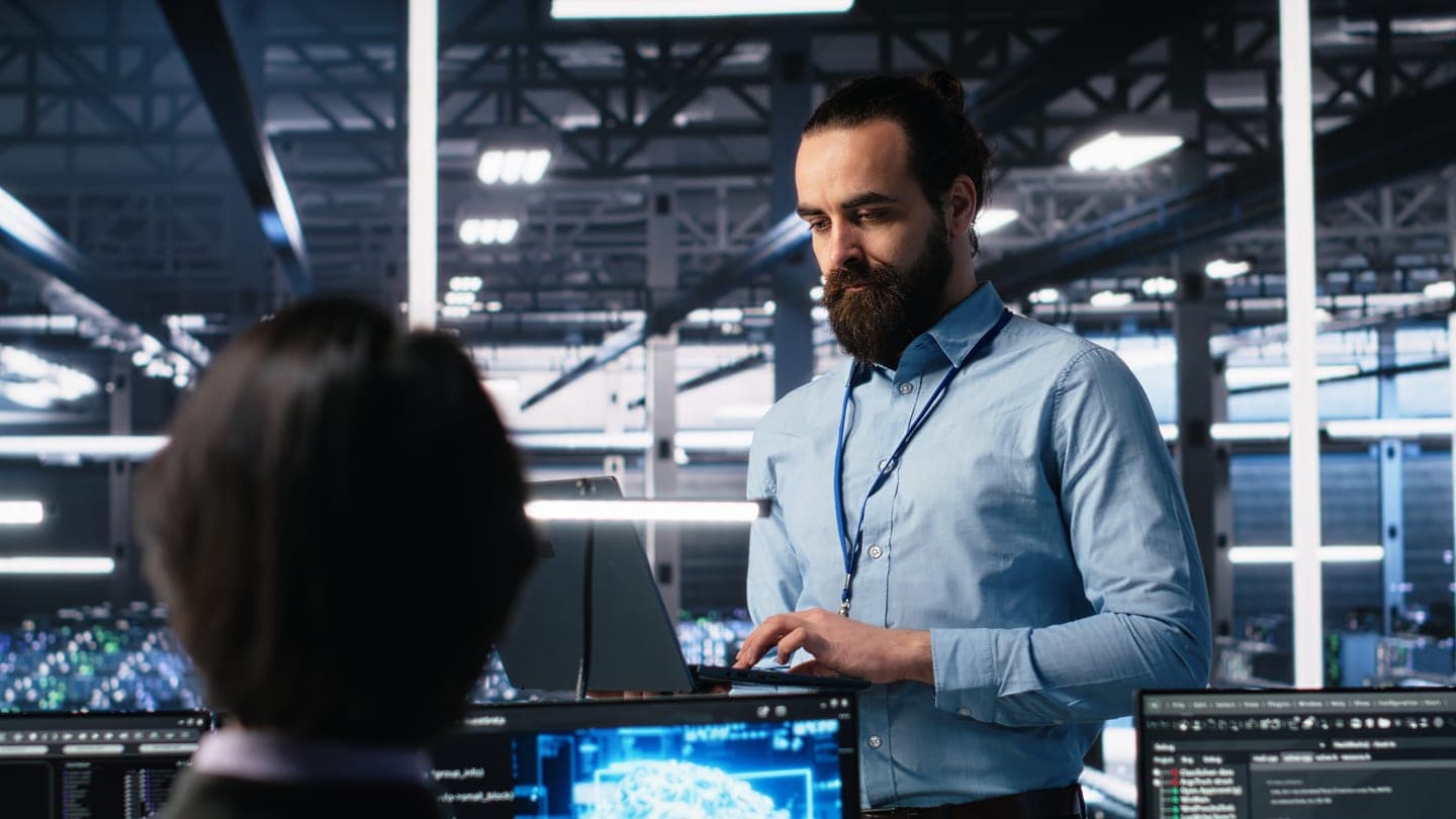 An engineer types on a laptop in a large server hall while a colleague faces monitors showing a glowing blue AI brain.