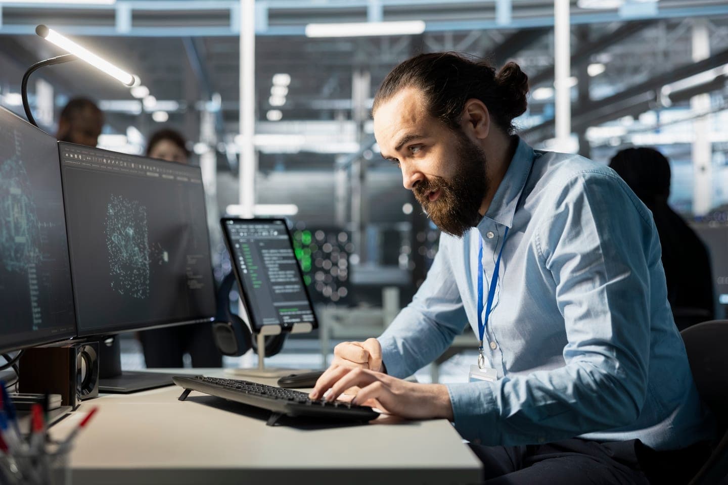 Bearded engineer in a light blue shirt typing at a desk with monitors showing neural network graphics, server racks lit behind him