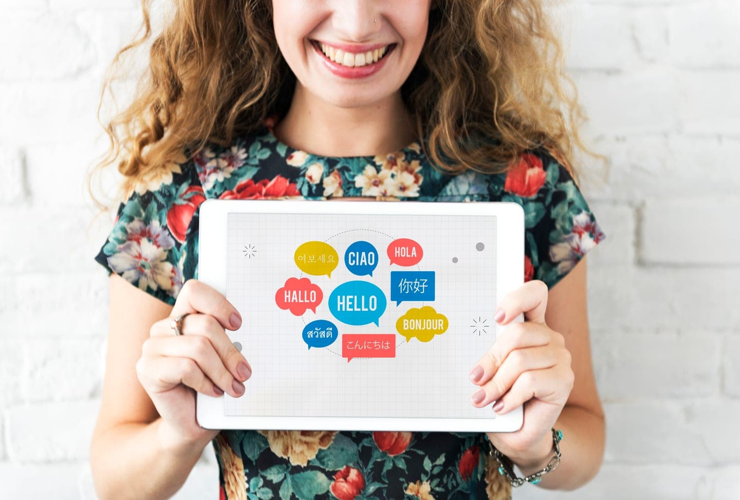 A smiling woman holds up a tablet showing coloured speech bubbles that say hello in several languages, including Hola, Ciao, Bonjour, Hallo and Chinese and Japanese greetings.