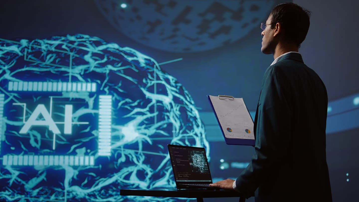A man in a dark suit holds a clipboard and checks a laptop at a standing desk while facing a large navy screen with a glowing cyan neural brain beside an AI chip.