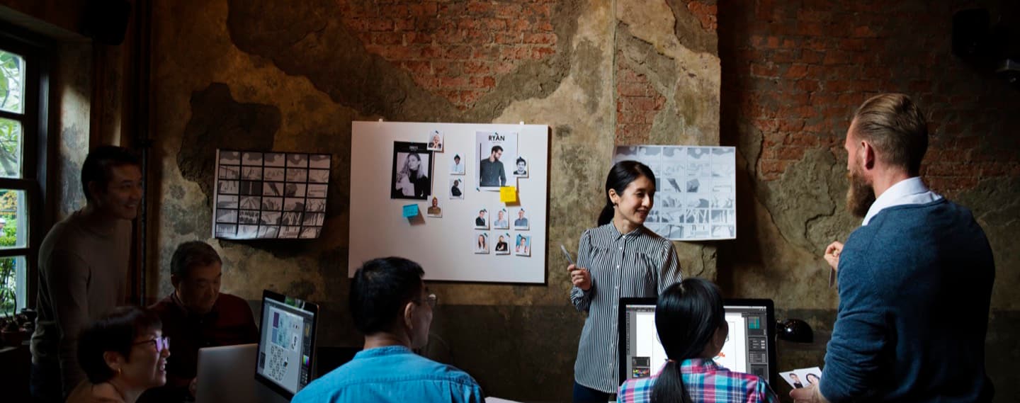 A team gathered around monitors in a brick-walled studio, discussing prints pinned to a large board.