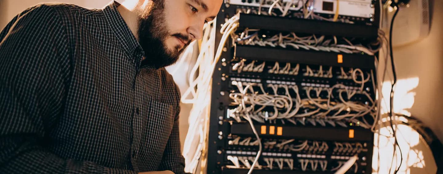 A bearded man in a checked shirt looks down at a laptop beside an open server rack packed with patch cables.