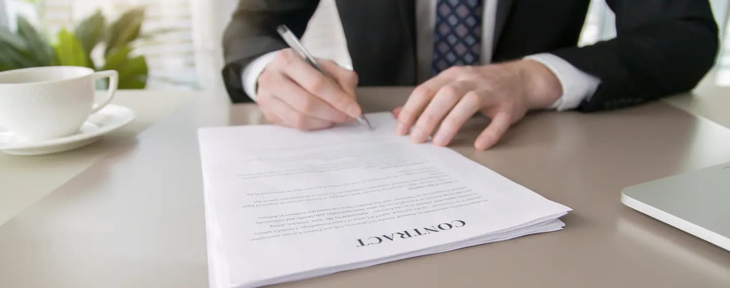 A businessman in a suit and tie signs a printed contract at a desk, with a coffee cup and the edge of a laptop either side of the document.