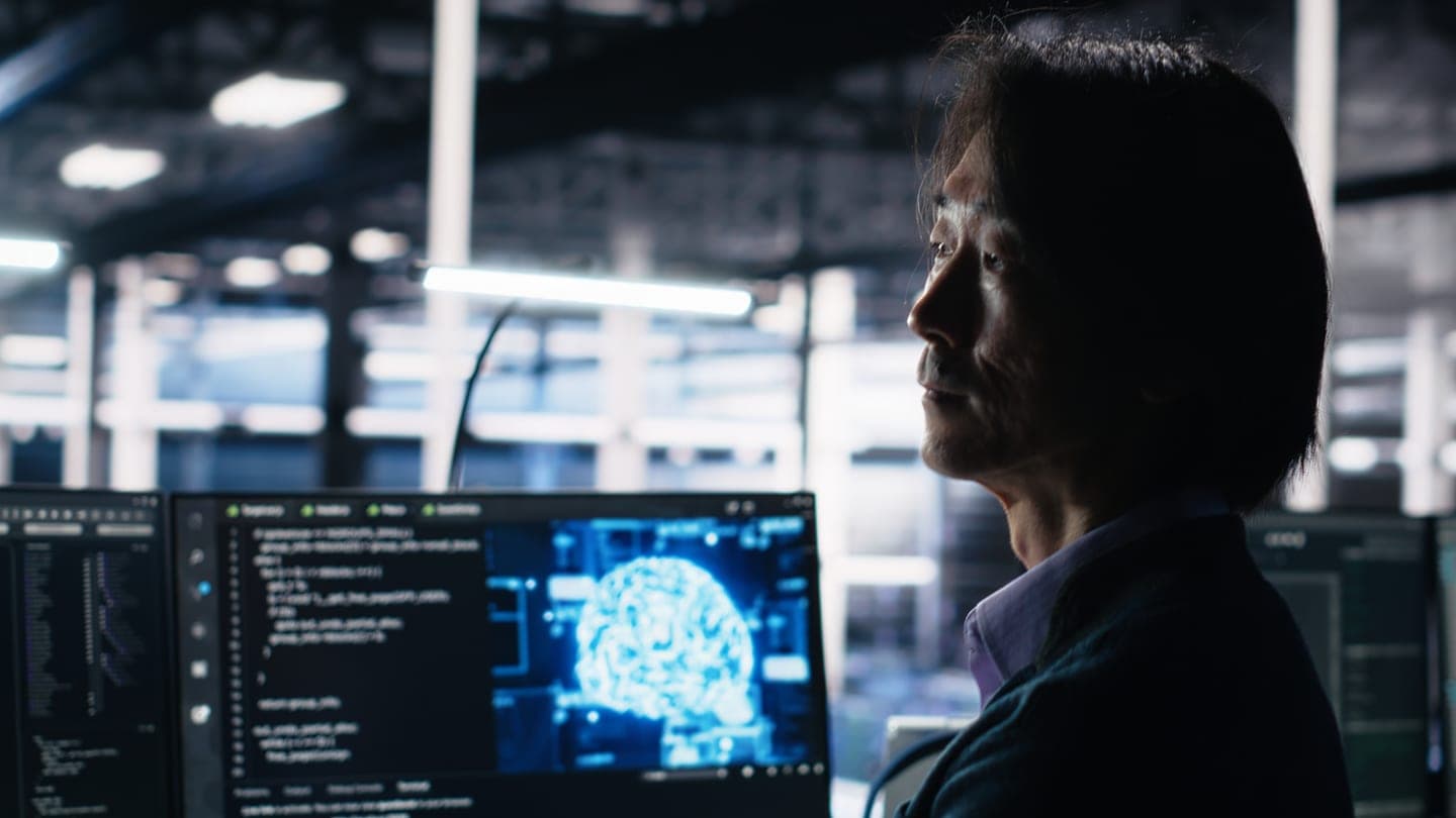 A man with shoulder-length dark hair sits in a dim, high-ceilinged server hall, looking past dark monitors that show lines of code and a glowing cyan neural network shaped like a brain.