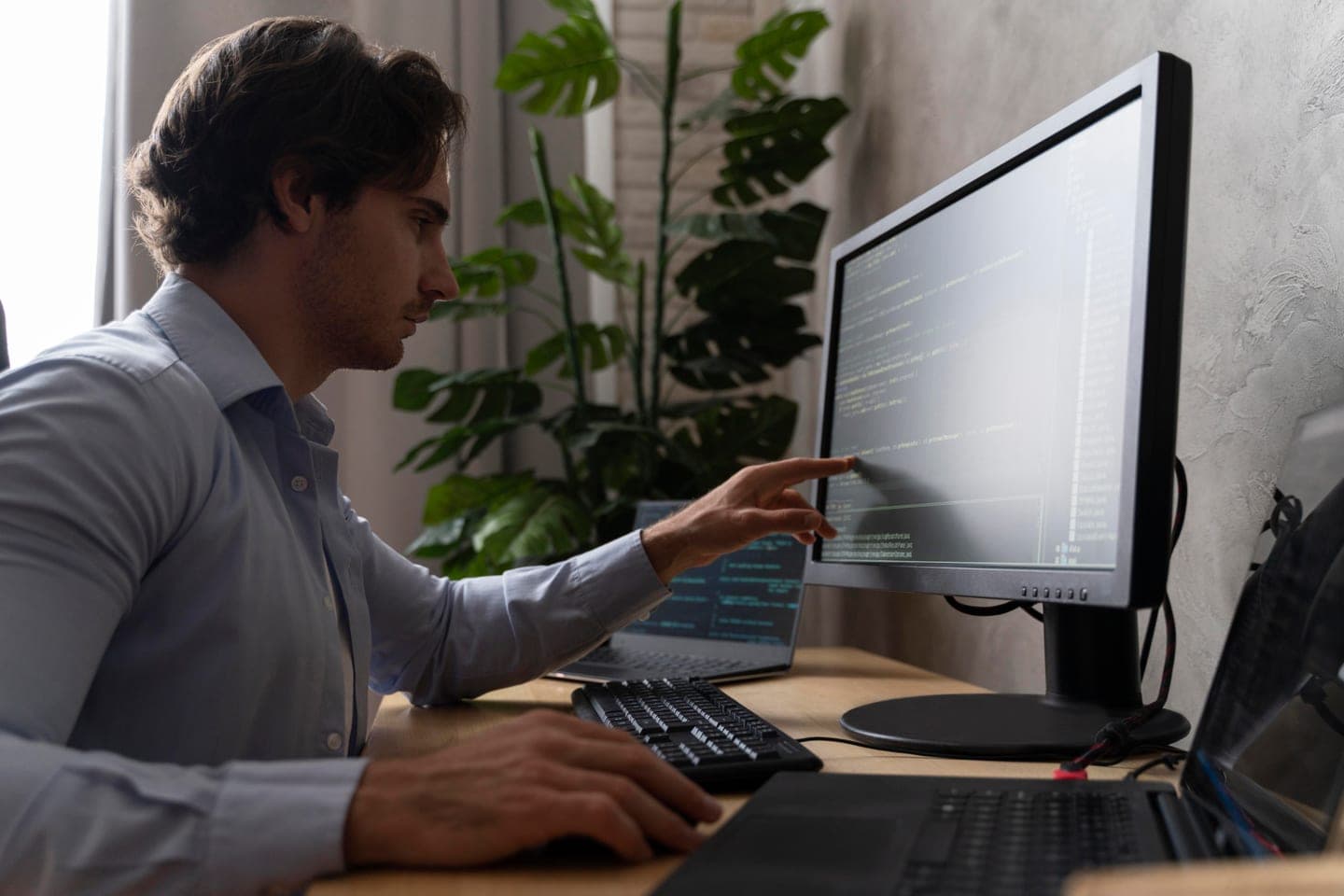 A man in a light blue shirt sits at a wooden desk beside a large leafy plant, pointing at lines of code on a dark monitor, with a laptop also showing code and a keyboard in front of him.