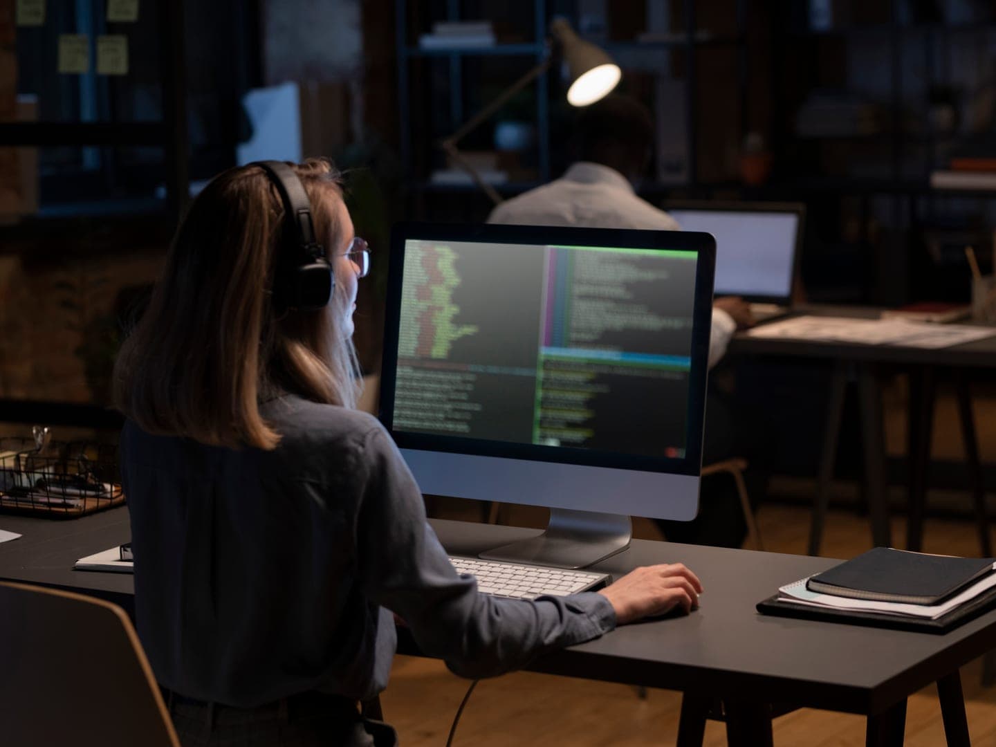 Developer in headphones at a desktop monitor showing coloured lines of source code in a dimly lit office, with a colleague at a laptop behind