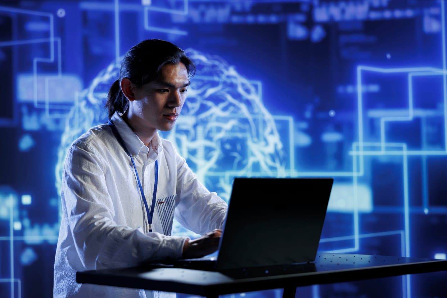 Engineer in a white shirt and lanyard typing on a laptop in front of a glowing blue wall of circuit lines and a holographic AI brain