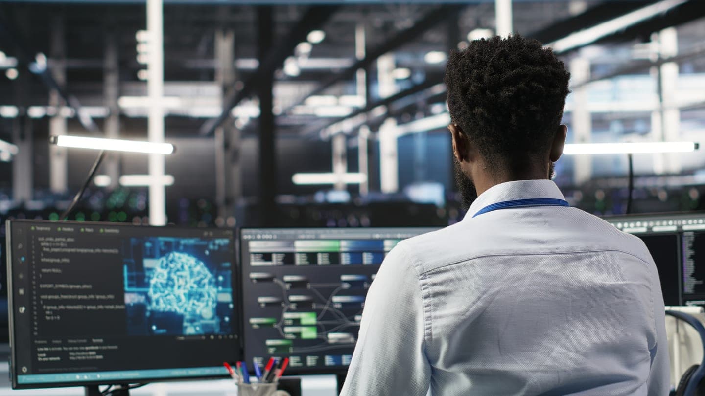 Engineer in a server hall facing two monitors that show code, a glowing neural network brain and a node graph of a machine learning pipeline