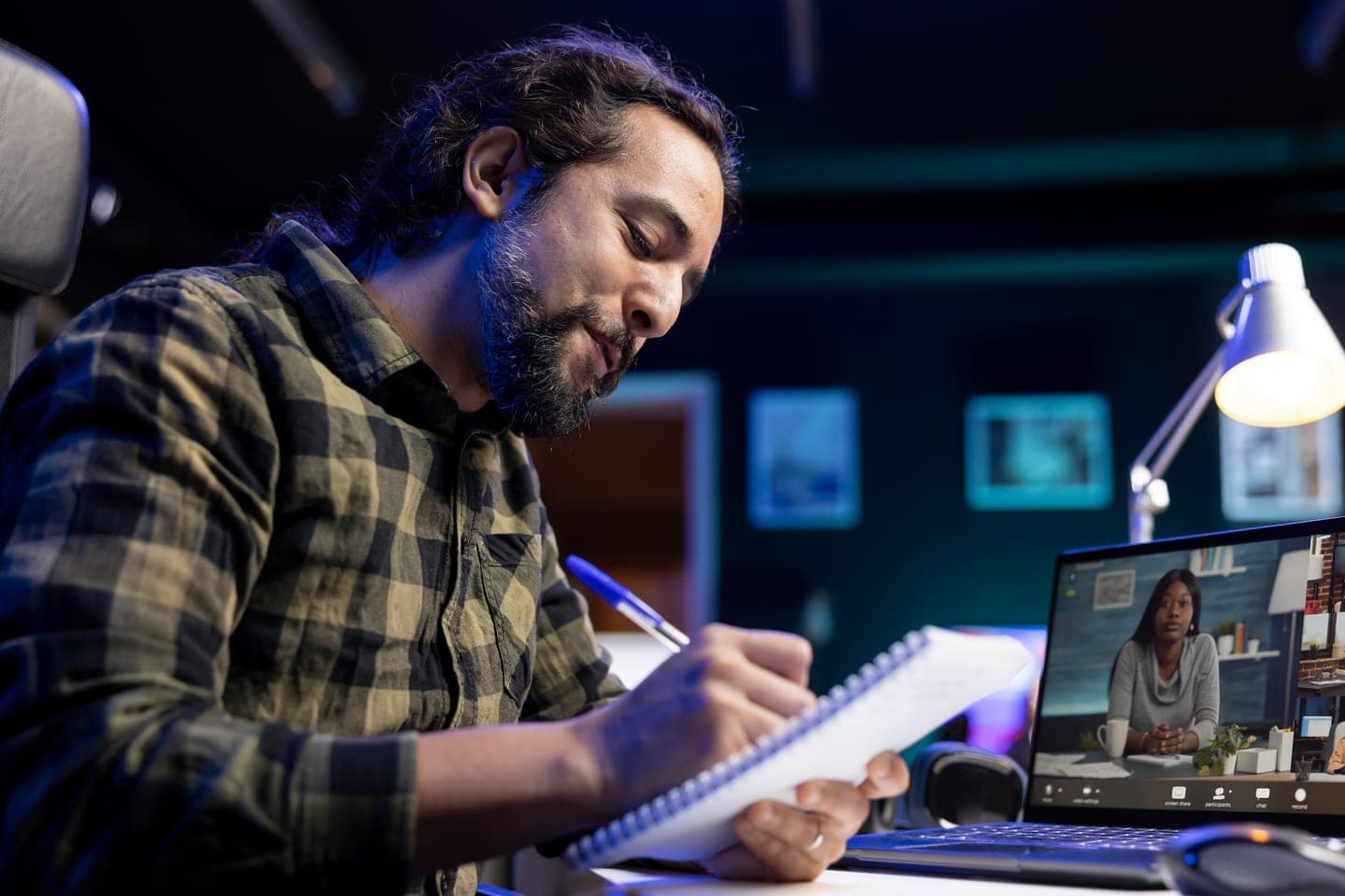 A bearded man in a checked shirt writes notes in a spiral notebook at a dark desk lit by a lamp, with a colleague on a video call on his laptop screen.