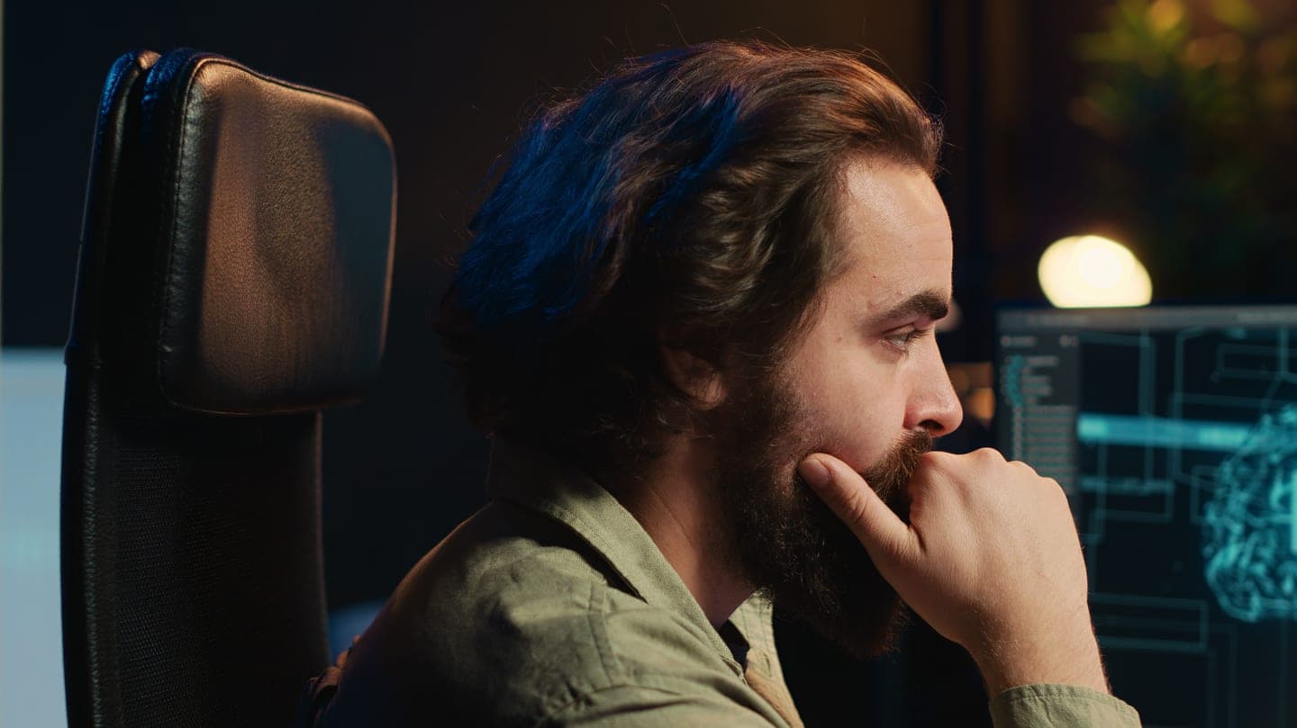 Bearded engineer in a dark office resting his chin on his hand as he studies a monitor showing a glowing cyan AI brain diagram and code